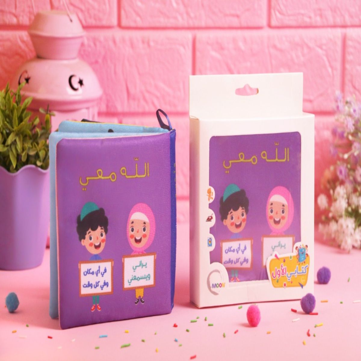 كتاب قماشي للأطفال بعنوان "الله معي" من سلسلة كتابي الأول، يظهر بجانب علبته الكرتونية بتصميم بنفسجي يغرس مراقبة الله في نفوس الصغار.