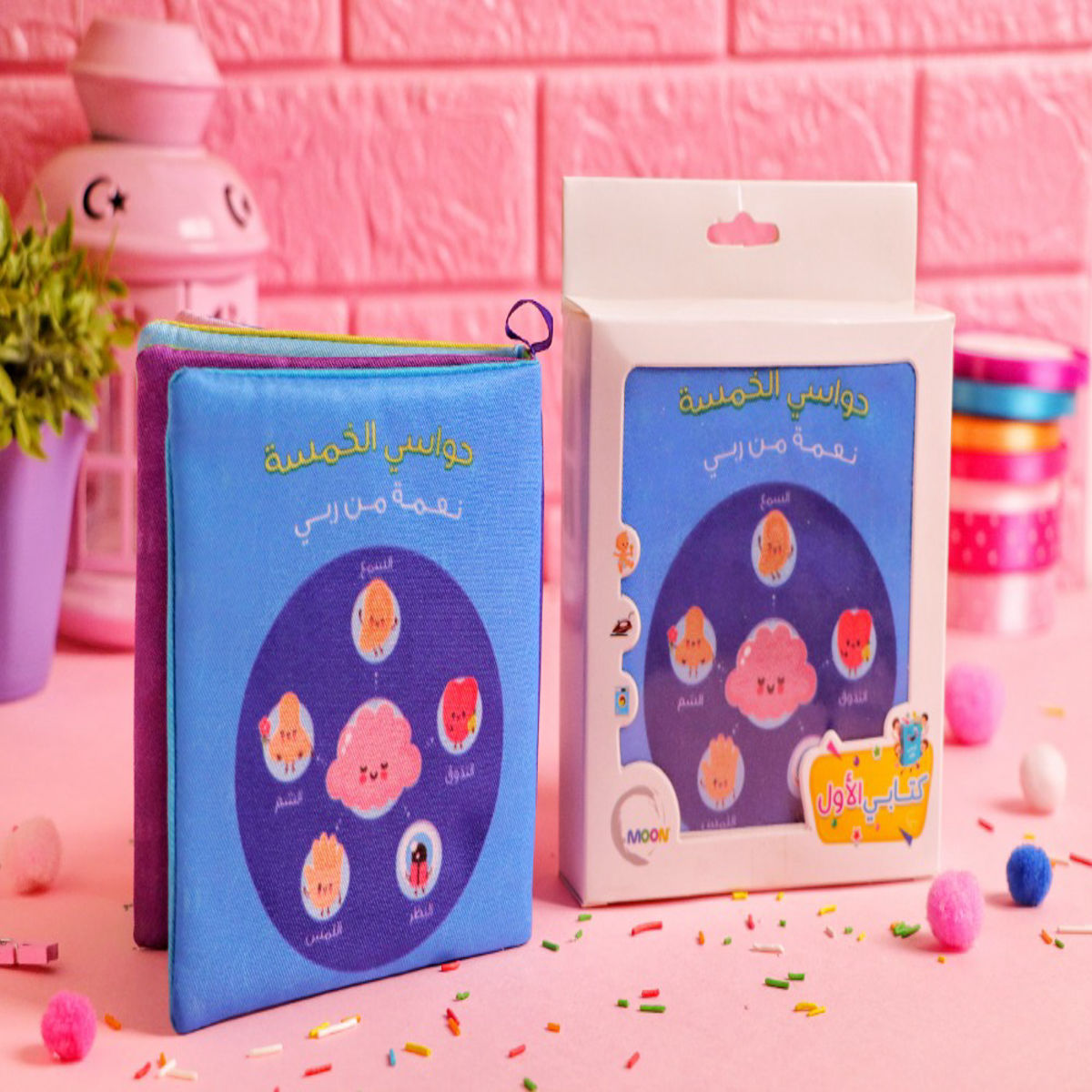 "كتاب قماش للأطفال بعنوان حواسي الخمسة نعمة من ربي بجانب علبة التغليف، يتميز برسومات كرتونية لطيفة للحواس، إصدار دار موون."