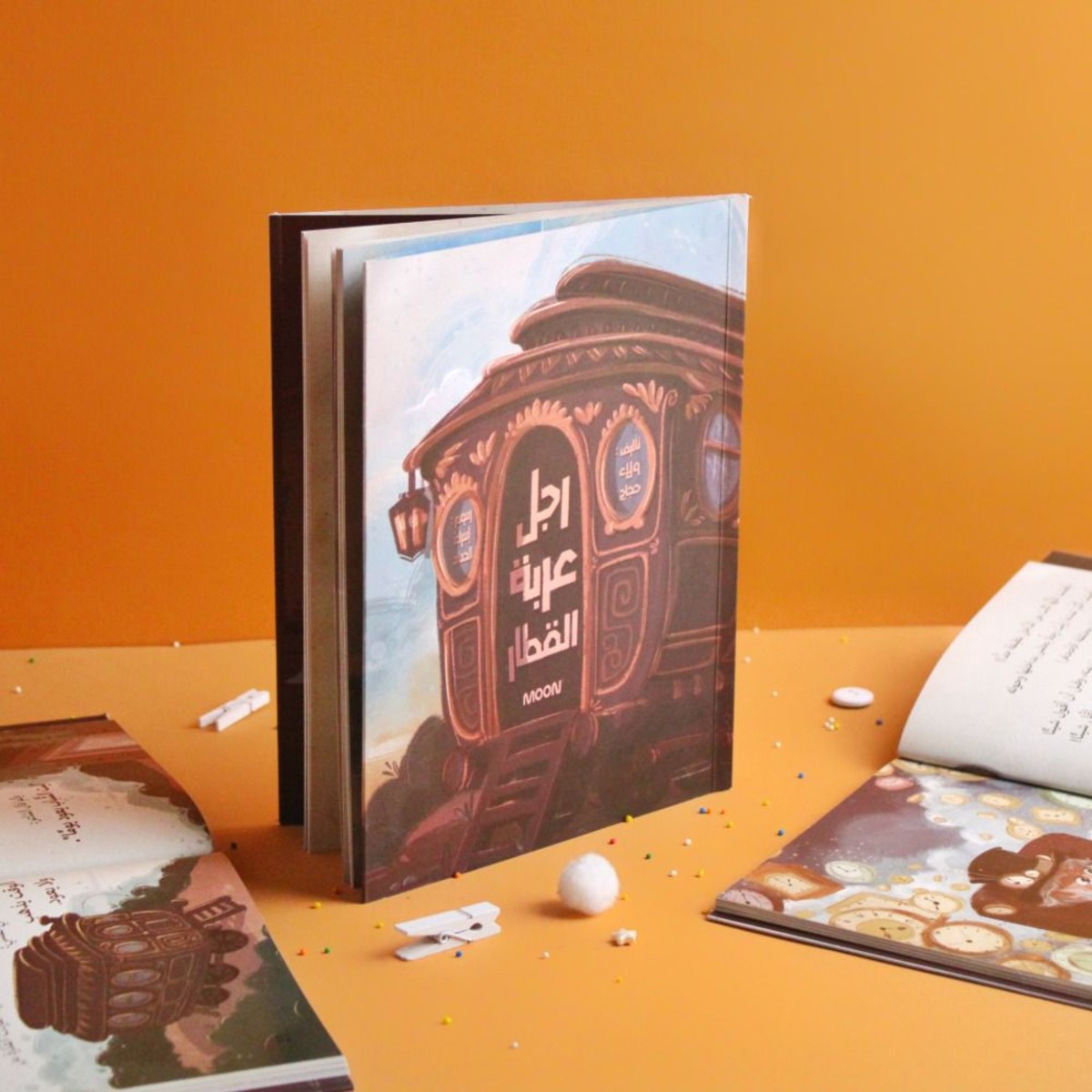 غلاف قصة الأطفال "رجل عربة القطار" من إصدارات MOON، يظهر طفلين ينظران من نافذة قطار إلى بائع متجول، قصة تعلم الأطفال الرحمة وقيمة العمل.